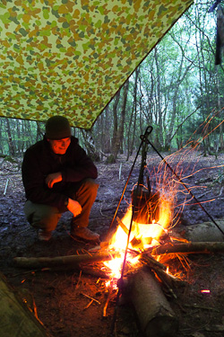 Campfire and tarp shelter in woods with man crouching