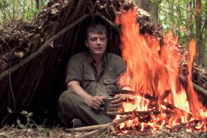 Frontier Bushcraft Online Bushcraft Courses banner - Paul Kirtley in an improvised shelter with fire in front