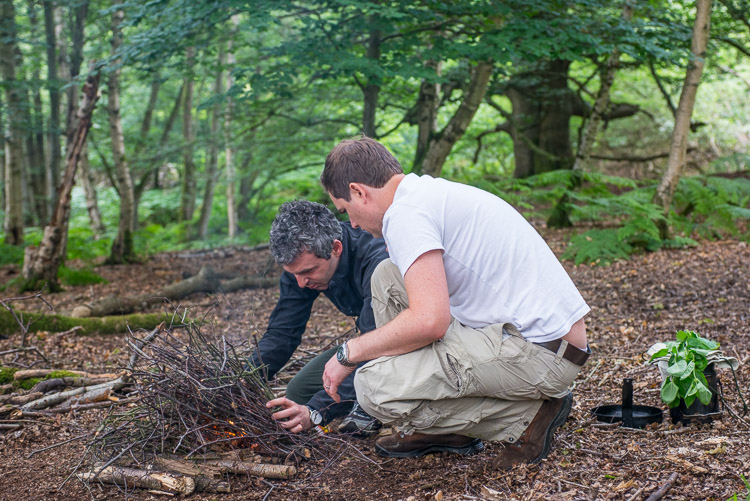 Two men , deep in the woods, lighting a fire, with water and wild herbs collected.