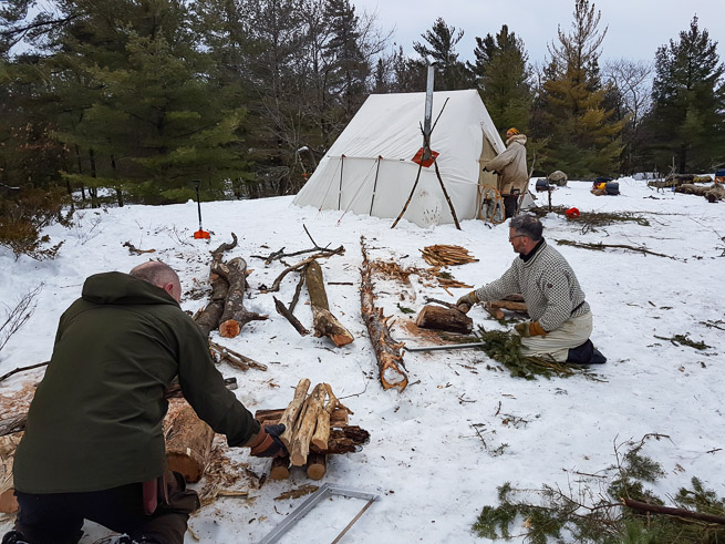 Men cutting wood in front of canvas winter tent with stove
