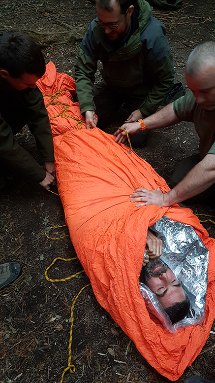 man with beard in foil blanket and orange fabric, with rope carrying improvisation