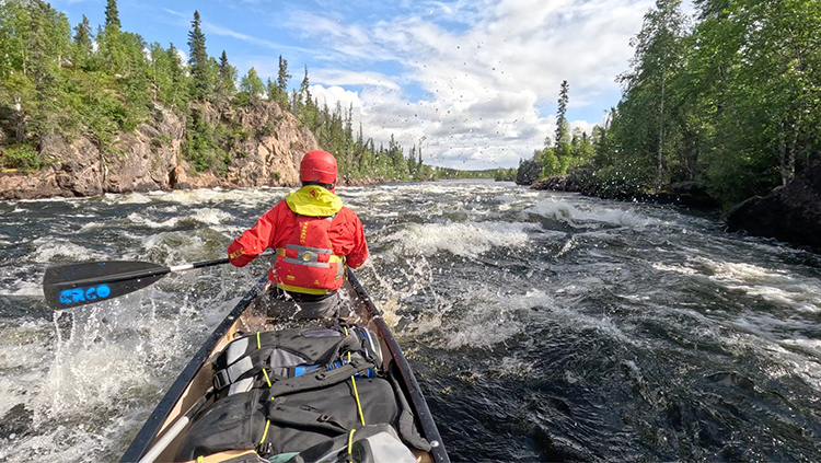 Stern paddler's view of his canoe, bow paddler and rocky, tree-lined boreal landscape as they enter a substantial whitewater rapid