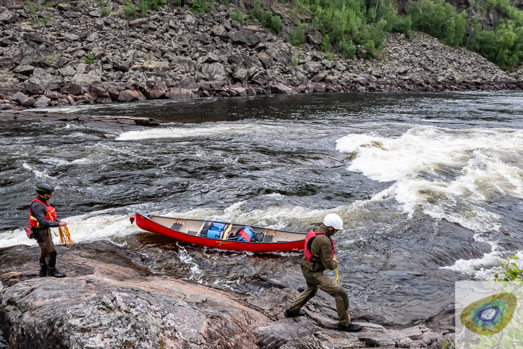 Two men with ropes controlling a canoe, walking quickly along a rocky riverbank, with whitewater rapids in the background